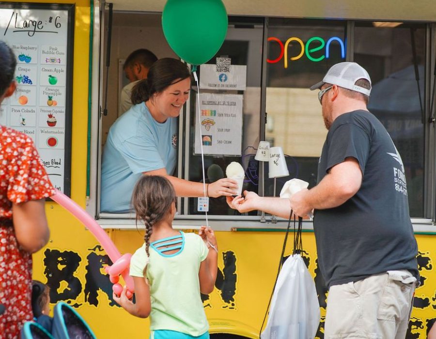 woman hands a snowcone to an adult dad who is standing next to a child holding a green balloon and holding a pink balloon sword