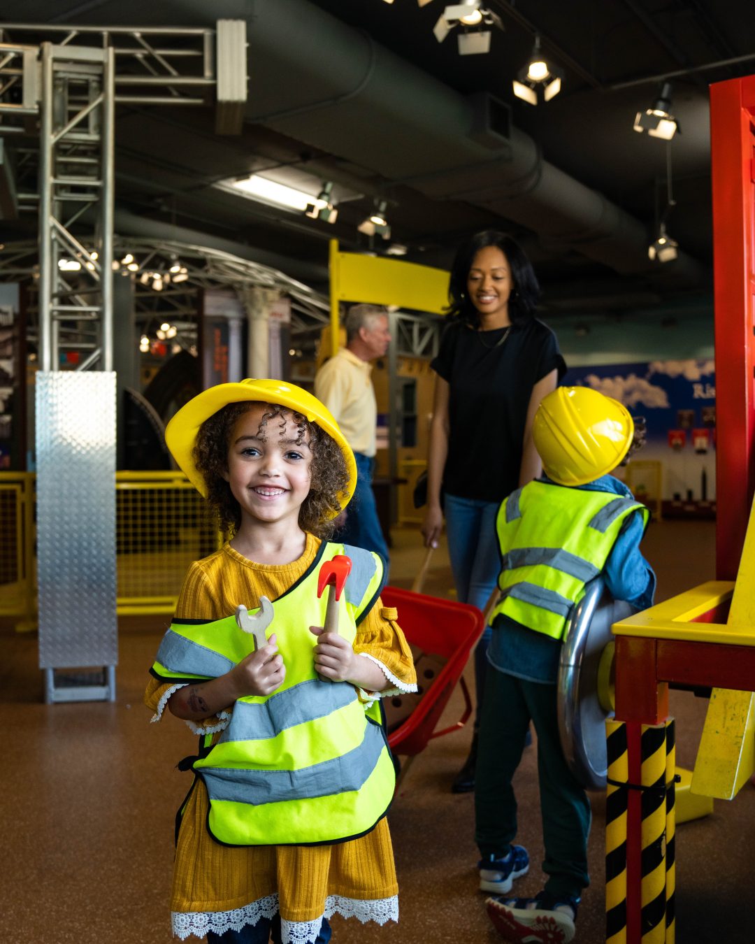 child smiles while wearing a construction vest and hard hat holding tools